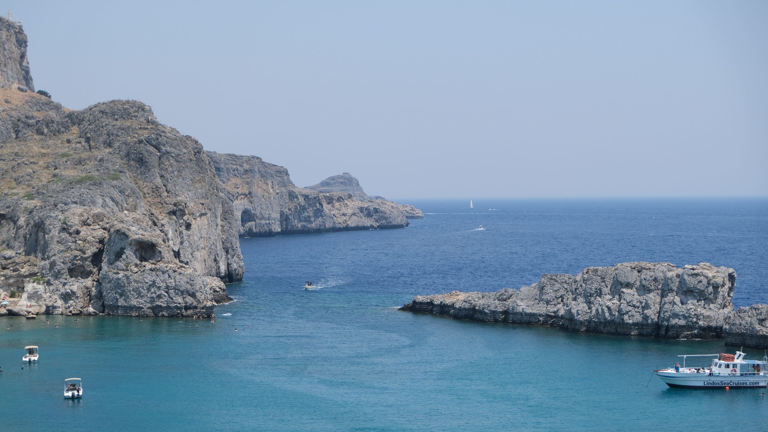 Serene view of Lindos coast with clear blue water, rocky cliffs, and boats floating under a clear sky.